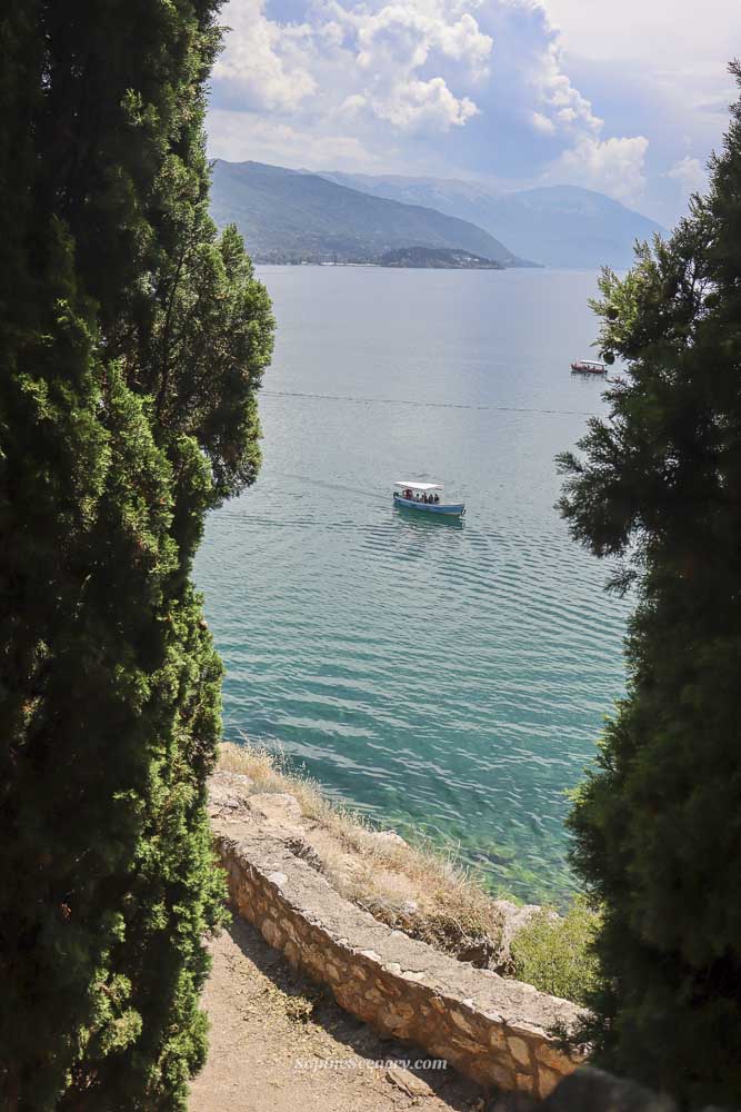 Blue water of Lake Ohrid between the trees you see a white boat on the lake. The sky and water a crystal clear blue and in the background you can see the mountains surrounding lake Ohrid in North-Macedonia