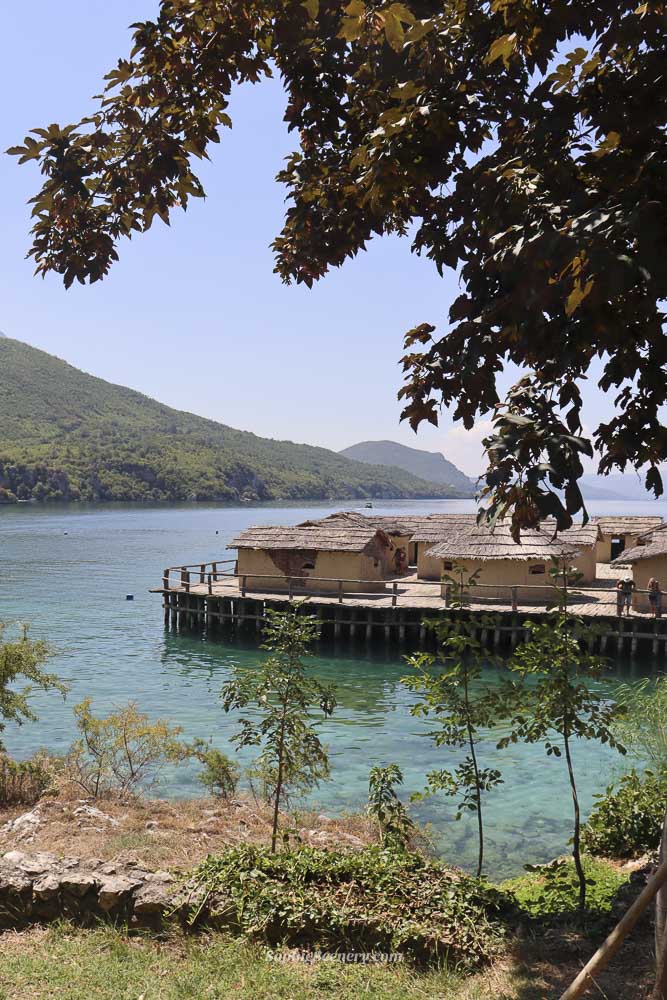 Bay of Bones open-air Museum on Lake Ohrid in North-Macedonia, showing reconstructed wooden houses built over turquoise water with mountains and greenery in the background on a sunny day