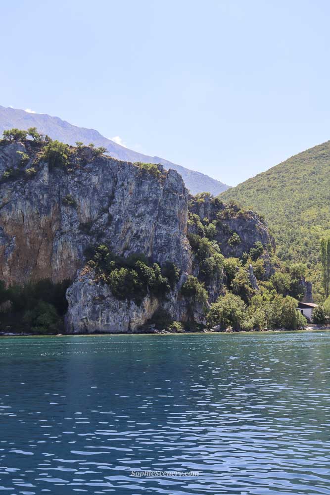 View of rocky mountain cliffs and green hills rising above deep blue water on Lake Ohrid, photographed from a boat on a calm sunny day in North Macedonia