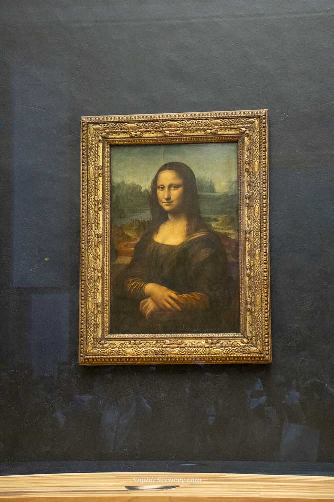 The Mona Lisa painting by Leonardo da Vinci displayed behind protective glass inside the Louvre Museum in Paris, surrounded by visitors.