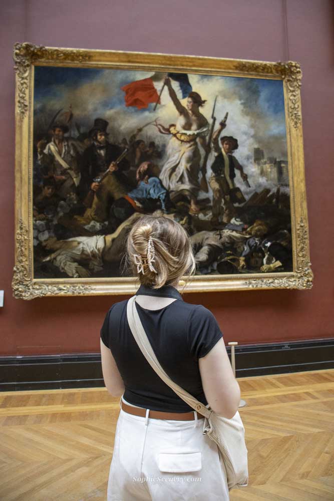 Young woman standing inside the Louvre Museum in Paris, viewing a famous French Revolution painting, highlighting free museum access for under-26 EU citizens.