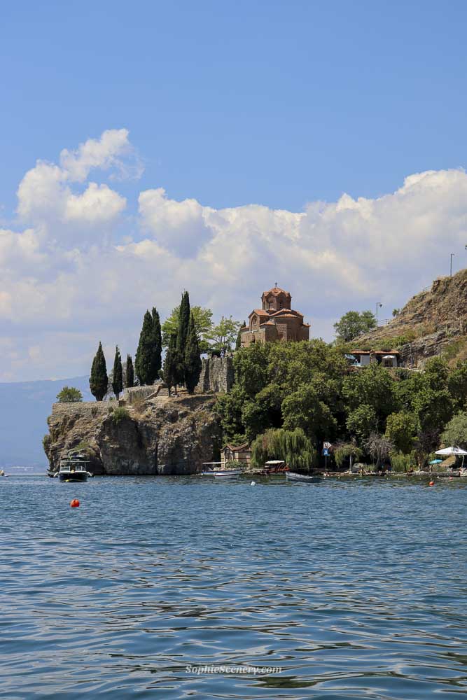 Church of Saint John the Theologian at Kaneo in Ohrid, North-Macedonia, perched on a cliff above Lake Ohrid with crystal-clear blue water and a bright blue sky in de background.