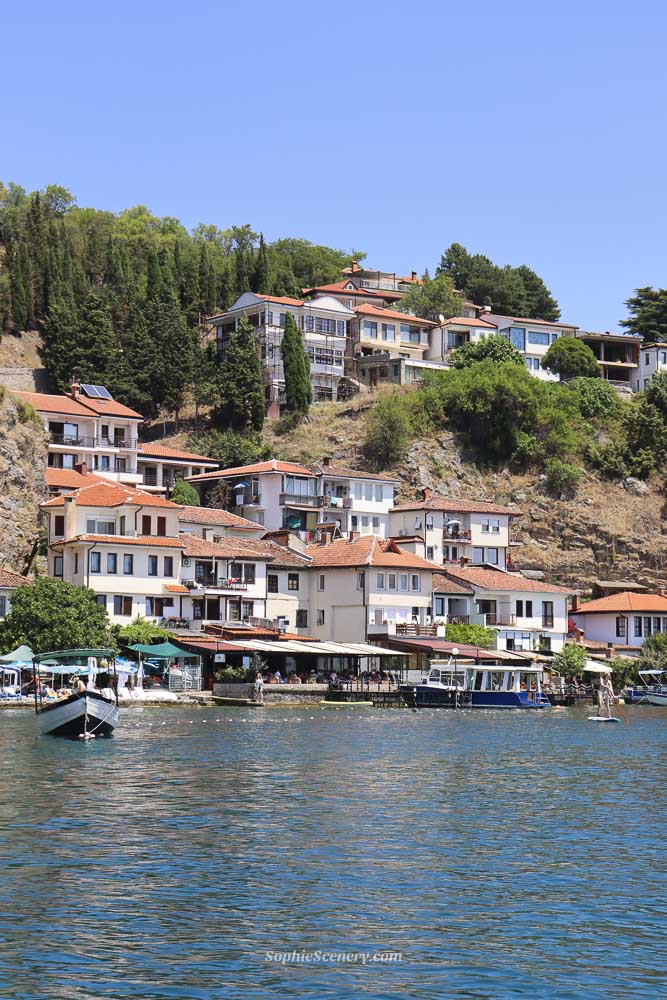 View of Ohrid from Lake Ohrid showing orange-roofed houses along the shoreline, calm blue water with small boats and a clear blue sky on a sunny day in North Macedonia