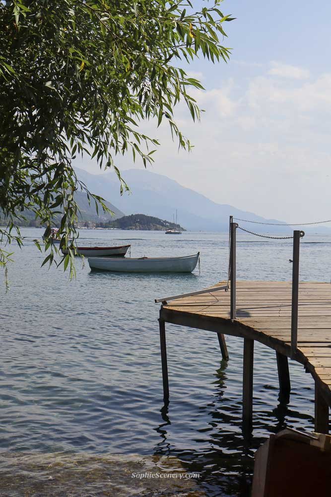 A watered wooden plank walkway leads through lush, overhanging green trees towards the bright blue waters of lake ohrid. In the distance a few people sit on the shore under the shade, with the 'bridge of wishes' path creating natural frame around the sunny beach scene in North Macedonia