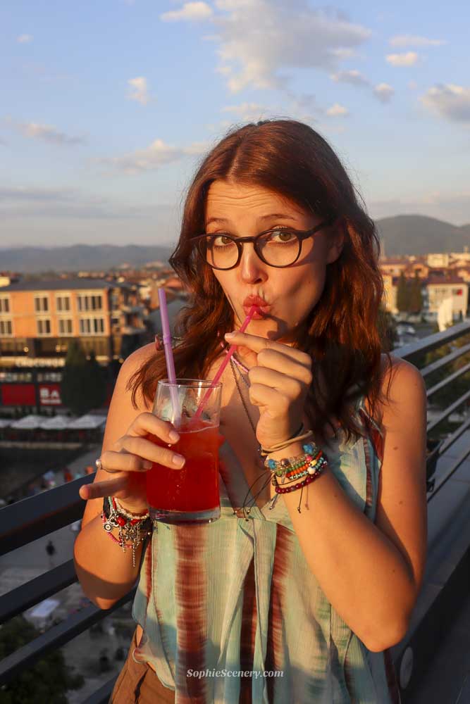 Woman drinking a cocktail on the Sky Drink rooftop in Strega, North Macedonia, during golden hour with city and mountain views in the background.