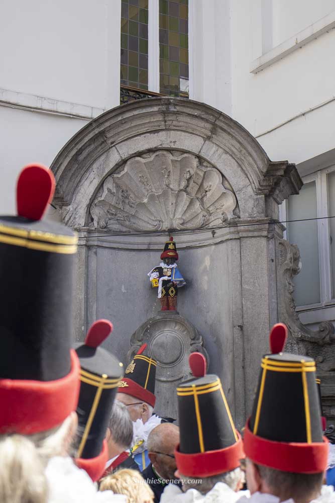 Manneken Pis statue in Brussels dressed in traditional ceremonial clothing during a local cultural celebration, with people around the statue wearing matching traditional outfits.