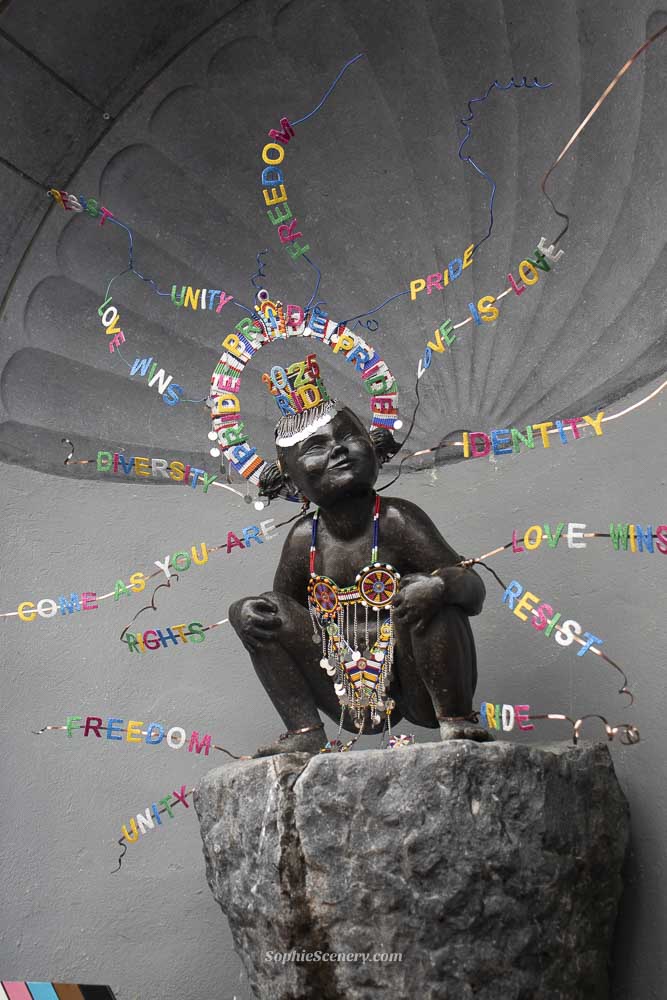 Jeanneke Pis statue in Brussels, Belgium, decorated with colorful Pride decorations in a city street setting.