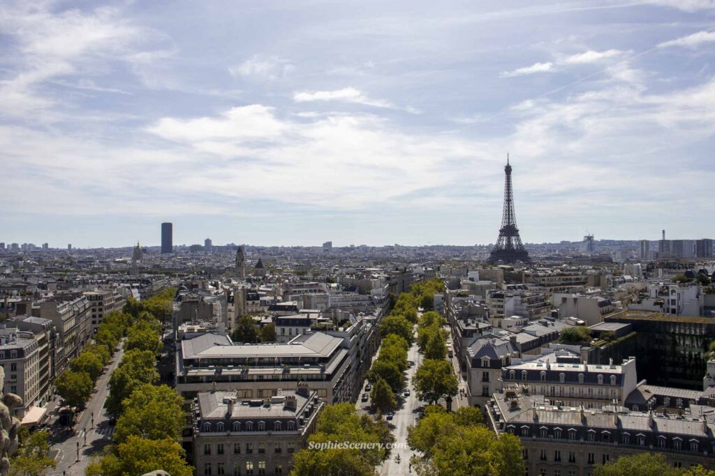 View from the top of the Arc de Triomphe in Paris with the Eiffel Tower visible in the distance, green trees lining the streets, and a bright summer sky.