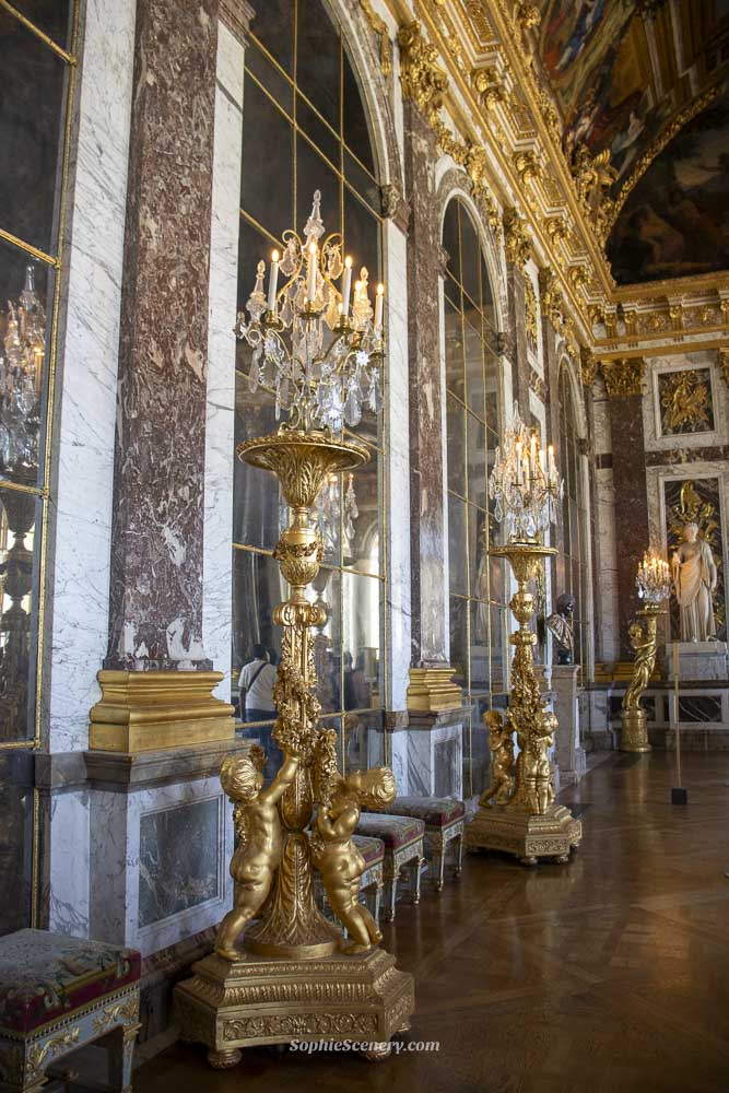 Classical marble statues displayed inside the Hall of Mirrors at the Palace of Versailles, reflecting in the mirrors under ornate chandeliers.