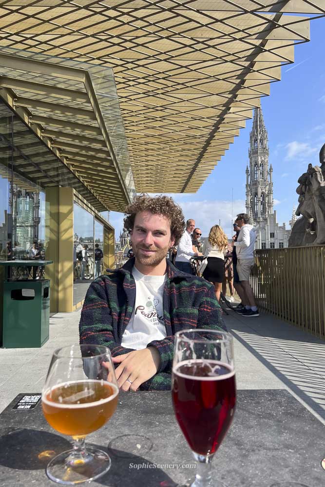 Boy sitting at a rooftop bar in Brussels with two Belgian beers on the table, overlooking historic city rooftops, Gothic church towers, and the Brussels skyline.