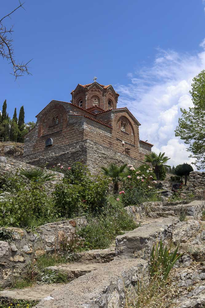 Church of Saint John the Theologian at Kaneo in Ohrid, North-Macedonia, perched on a cliff above Lake Ohrid with crystal-clear blue water and a bright blue sky in de background.