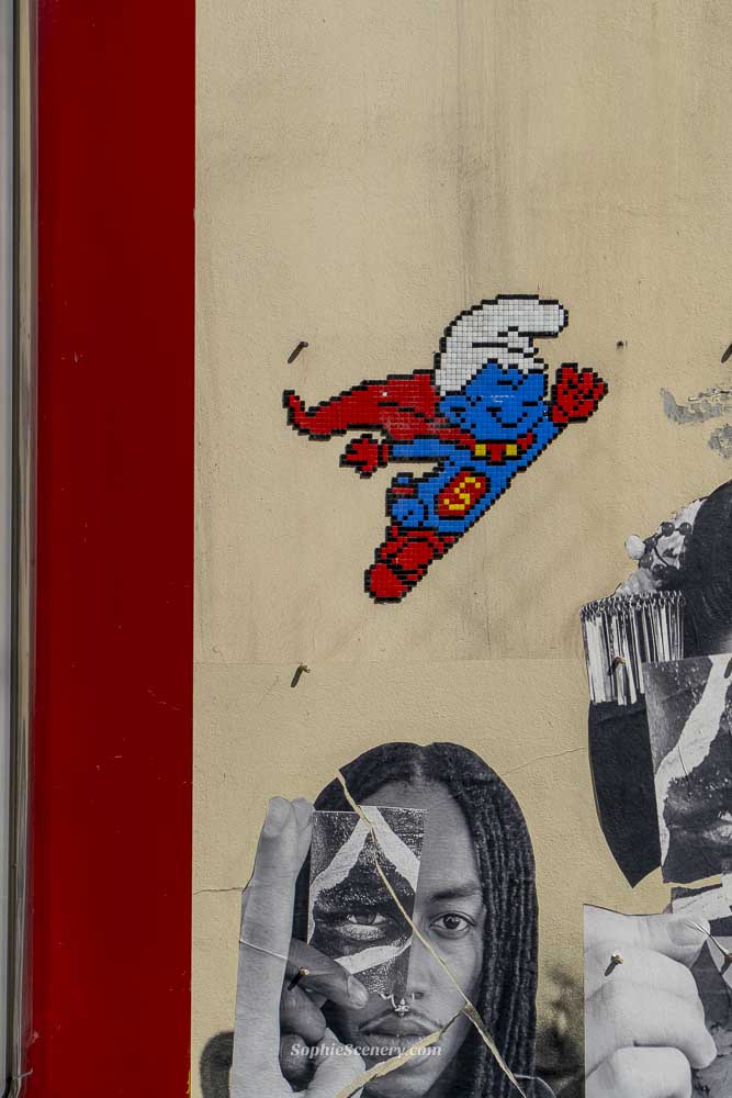 Pixel-style Super Smurf wall art on a building in Brussels, Belgium, featuring the beloved blue comic character dressed as a superhero with a red cape. The artwork is part of Brussels’ famous Comic Strip Route and is displayed above a black-and-white mural on a city wall.