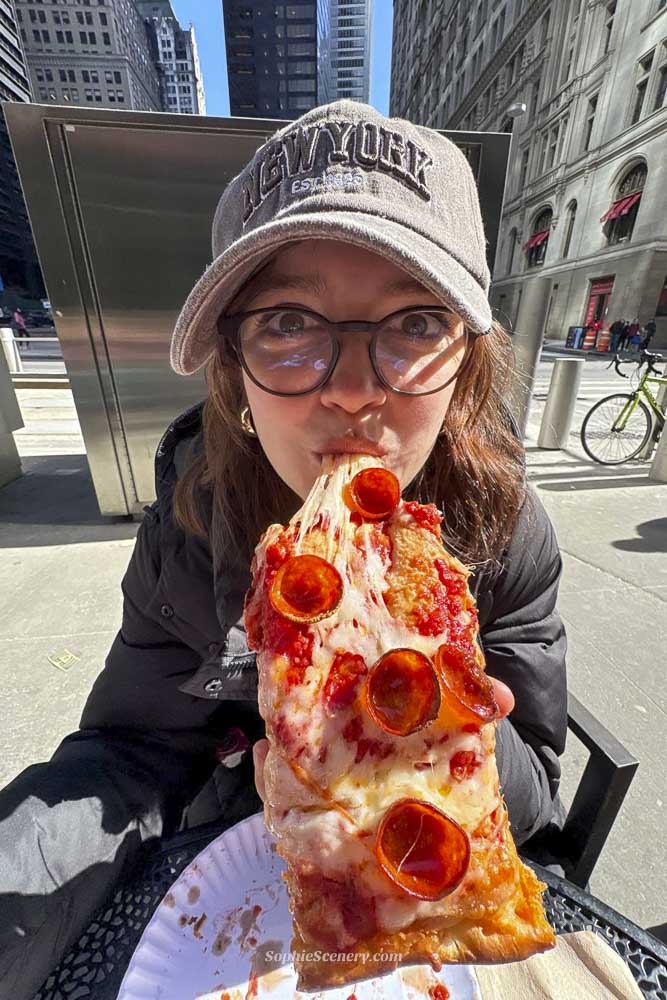 A person wearing glasses and a gray New York baseball cap sits outdoors in a city setting, holding and eating an oversized slice of pepperoni pizza with melted cheese stretching toward their mouth. Tall buildings and a bicycle are visible in the background on a sunny day.