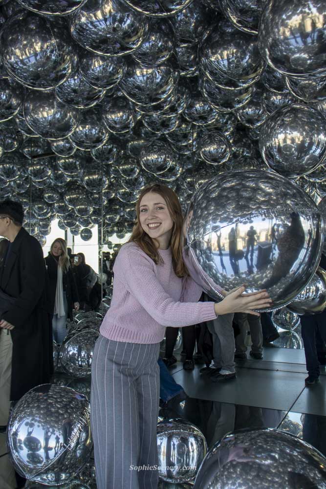 SUMMIT One Vanderbilt NYC, silver balloon room Visitor inside the mirrored balloon installation at SUMMIT One Vanderbilt in New York City, surrounded by reflective silver spheres and glass mirrors creating immersive skyline reflections.