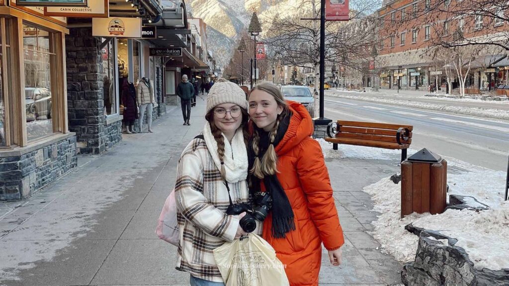 Two girls standing on Banff Avenue in Banff, Canada during winter, with snowy mountains in the background. One girl is wearing a bright orange winter coat while the other wears a cream plaid jacket and white beanie. The photo captures the cozy winter atmosphere of Banff National Park and a girls trip to Lake Louise in Canada.