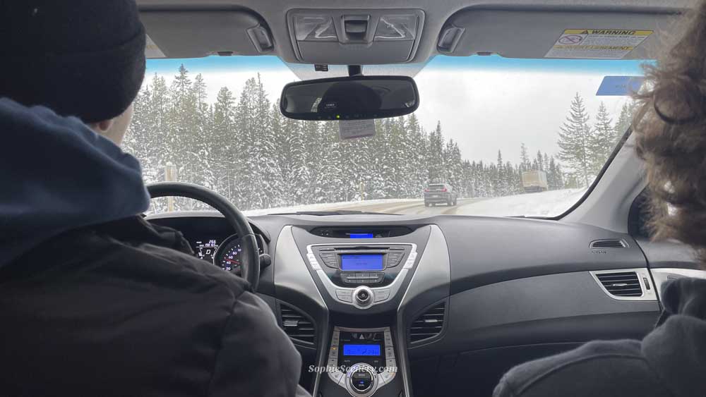 Unexpected Road Trip to Lake Louise View from inside a car driving through Banff National Park during winter, with snowy mountains visible through the window. Two boys are sitting in the front seats while the road trip toward Lake Louise captures a cozy and adventurous winter travel moment in Canada.