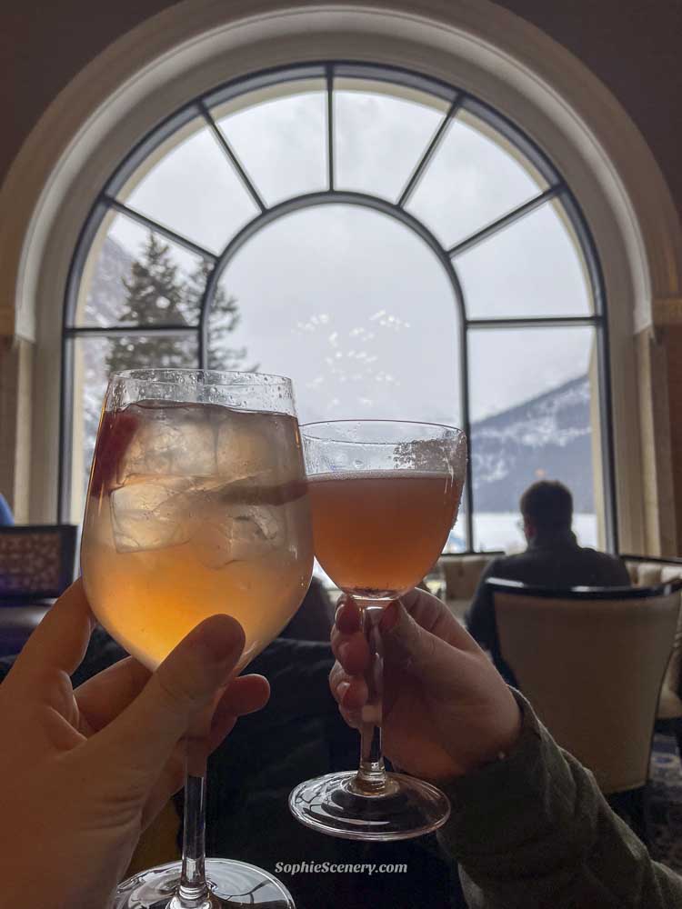 Cocktails at Fairmont Chateau Lake Louise Two cocktails being held inside the Fairmont Chateau Lake Louise hotel with a large arched window overlooking snowy mountains and frozen Lake Louise in Banff National Park during winter. The cozy atmosphere captures a luxurious winter travel experience in Canada.
