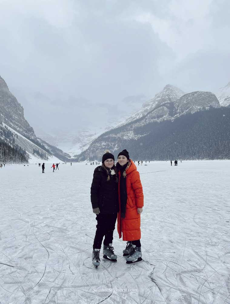 Ice Skating on Lake Louise in Winter | Banff Travel Guide Two girls ice skating on the frozen Lake Louise in Banff National Park during winter, surrounded by snowy mountains and a cloudy sky. One girl is wearing a bright orange winter coat while both stand on the ice with skates, capturing a magical winter travel moment in Canada.